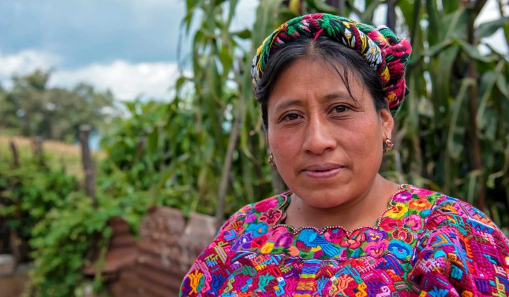 Close-up of an indigenous Guatemalan woman standing in natural setting with green trees and foliage behind her.