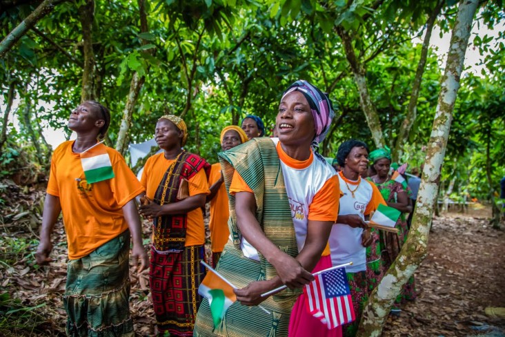 Women singing in a cocoa farm holding the American and Ivoirian flags.