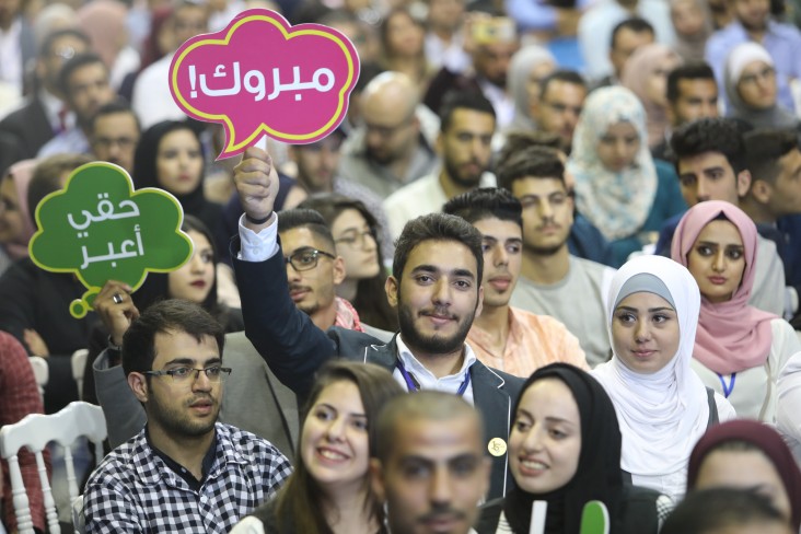 A young man holds up a sign and smiles in a group of youth at a graduation for a civic education program.