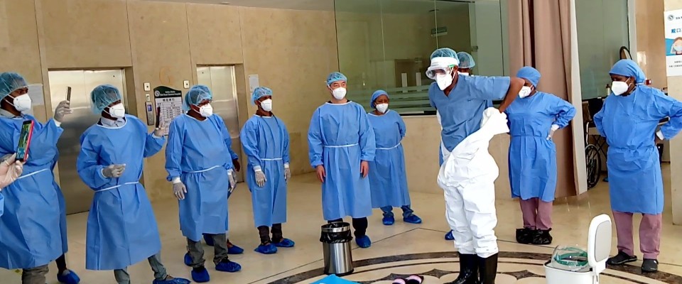 A group of people in medical scrubs and face masks stands in a circle around a person putting on hazmat clothing