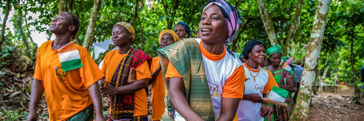 Women singing and dancing in a cocoa farm holding the American and Ivoirian flags.