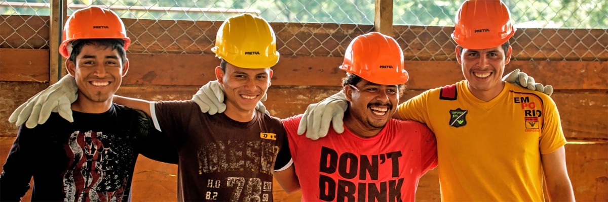 Four smiling craftsmen wearing hard hats and gloves stand in a row with arms across each others' shoulders